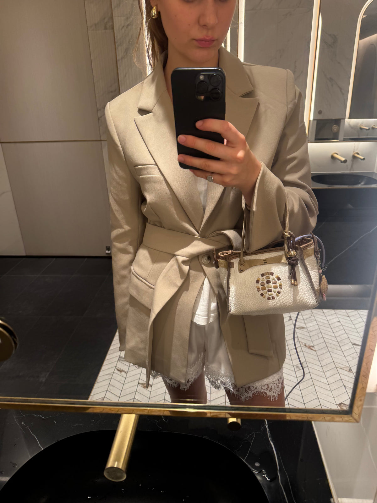 A woman takes a mirror selfie in a bathroom while wearing a beige tailored blazer with a matching belt, holding a black smartphone and carrying a starbell  handbag.